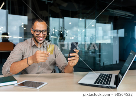 Smiling young African American man sitting in office at desk with laptop. He is holding a credit card, using the phone. Smiling young African American man sitting in office at desk with laptop. He is holding a credit card, using the phone. 98564522