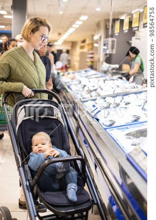 Casualy dressed mother choosing fish in the fish market department of supermarket grocery store with her infant baby boy child in stroller Casualy dressed mother choosing fish in the fish market department of supermarket grocery store with her infant baby boy child in stroller 98564758