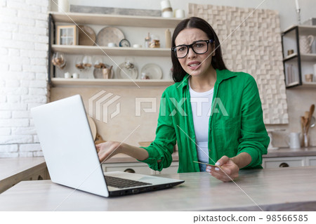 Angry young woman sitting at home in the kitchen, working on a laptop, having problems with a credit card, bill, payment. He spreads his hands and looks at the camera in confusion. Angry young woman sitting at home in the kitchen, working on a laptop, having problems with a credit card, bill, payment. He spreads his hands and looks at the camera in confusion. 98566585