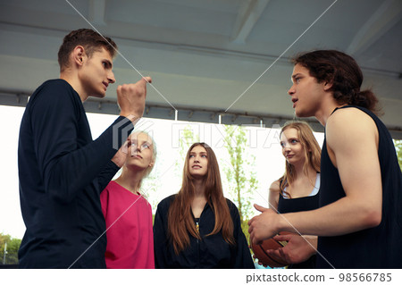 A group of cheerful young people girls and guys are discussing and debating while playing basketball on a street playground. Diversity men and women play basketball together 98566785