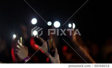 Fans Raise Hands and takes a photos in Front of Bright Colorful Strobing Lights on Stage. 98568018