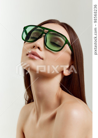Enjoyment. Portrait of young beautiful girl with smooth healthy skin posing in green sunglasses over grey studio background. Concept of beauty, fashion Enjoyment. Portrait of young beautiful girl with smooth healthy skin posing in green sunglasses over grey studio background. Concept of beauty, fashion 98568236