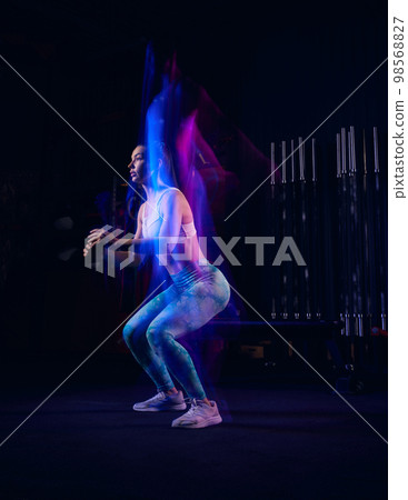 Portrait of muscular strong sportive young girl doing exercises, workout at dark gym in mixed neon light. Active and healthy lifestyle, sport, fitness concept Portrait of muscular strong sportive young girl doing exercises, workout at dark gym in mixed neon light. Active and healthy lifestyle, sport, fitness concept 98568827