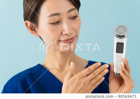 一位穿著磨砂膏並解釋面部按摩器的年輕女性美容形象 一位穿著磨砂膏並解釋面部按摩器的年輕女性美容形象 98569471