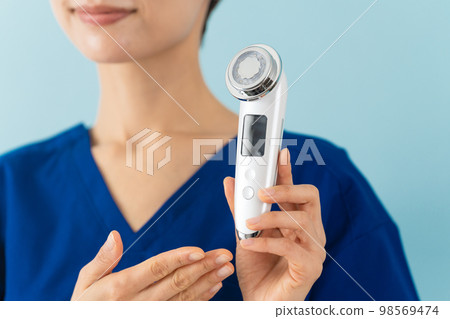 一位穿著磨砂膏並解釋面部按摩器的年輕女性美容形象 一位穿著磨砂膏並解釋面部按摩器的年輕女性美容形象 98569474