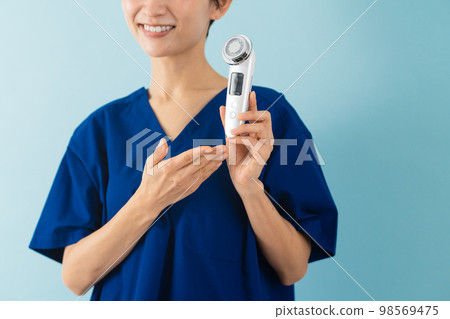 一位穿著磨砂膏並解釋面部按摩器的年輕女性美容形象 一位穿著磨砂膏並解釋面部按摩器的年輕女性美容形象 98569475