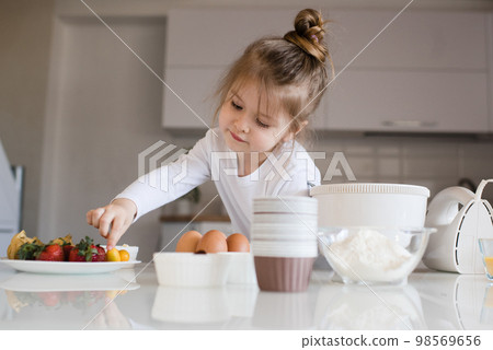 Smiling baby girl 3-4 year old making dough in white bowl for cake on kitchen table at home close up. Kid cook pastry with ingredients. Childhood. 98569656