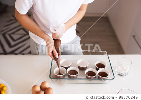 Woman making chocolate muffins at home. Put batter to cupcake forms in glass baking tray on kitchen table close up. 98570249