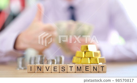 Word investment on cubes, stacks of coins and gold bars and investor with thumbs up in background. Word investment on cubes, stacks of coins and gold bars and investor with thumbs up in background. 98570817