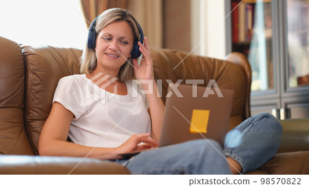 Woman with headphones smiling and looking at laptop screen while sitting on the couch. Woman with headphones smiling and looking at laptop screen while sitting on the couch. 98570822