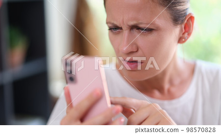 Woman with serious face looks doubtful at smartphone screen. . Woman with serious face looks doubtful at smartphone screen. . 98570828