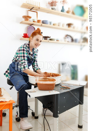 Cheerful female sculptor while she making clay pot on pottery wheel. Idea of small business and entrepreneurship. Home hobby, entertainment and leisure. Art studio 98571005