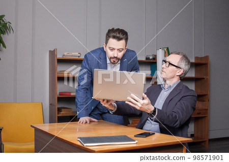 Mature businessman in blue suit is showing work on laptop to his colleague in office 98571901