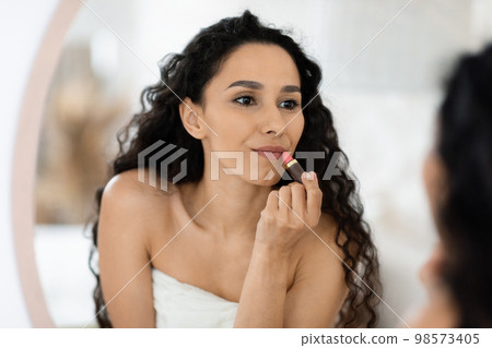 Glad pretty islamic millennial curly female in towel applying lipstick on lips, looking in mirror in bedroom Glad pretty islamic millennial curly female in towel applying lipstick on lips, looking in mirror in bedroom 98573405