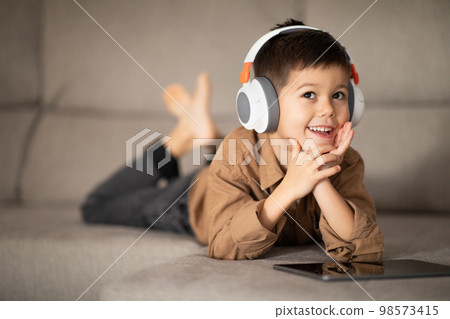 Happy pensive little boy in wireless headphones thinks, creative idea, watching video on tablet, lies on sofa Happy pensive little boy in wireless headphones thinks, creative idea, watching video on tablet, lies on sofa 98573415