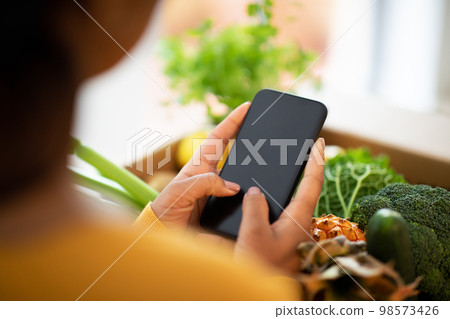 Millennial african american woman unpacks cardboard box with organic vegetables uses smartphone with blank screen Millennial african american woman unpacks cardboard box with organic vegetables uses smartphone with blank screen 98573426