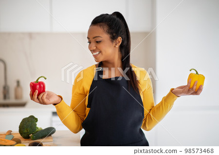 Cheerful millennial african american female in apron chooses peppers in hands at table with vegetables Cheerful millennial african american female in apron chooses peppers in hands at table with vegetables 98573468
