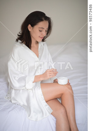 Happy young european woman in bathrobe with perfect legs sits on white bed with jar of cream in bedroom 98573486