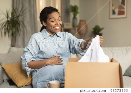 Happy contented pregnant african american lady unpacking box and holding clothes, sitting on couch and touching belly 98573563