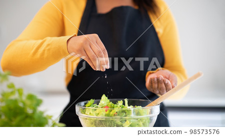 Millennial african american woman in apron salting salad, cooking dinner at table with organic fresh vegetables 98573576