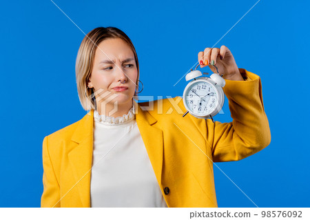 Irritated woman holding alarm clock in hand. Blue background. Early 6 o'clock Irritated woman holding alarm clock in hand. Blue background. Early 6 o'clock 98576092