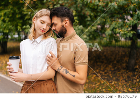 Front view of pretty young couple in love standing embracing with closed eyes at city park on background of trees. Bearded tattooed man and charming woman walking spending time together on summer day. 98576179