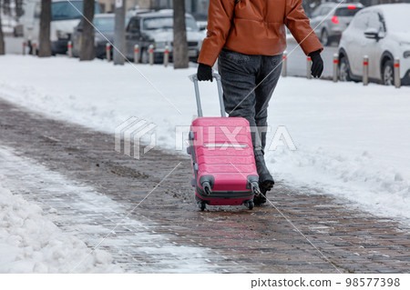 A traveler woman walks along a paved sidewalk in the winter season, carrying a travel bag on wheels. Copy space. 98577398