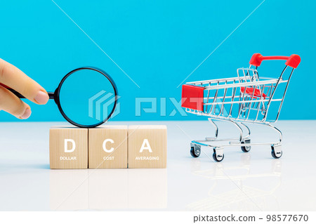 Magnifying glass in woman's hand and DCA word on a wooden cube in idea Dollar Cost Averaging investment strategy near empty shopping cart. Saving stock or savings on a monthly, quarterly basis. 98577670