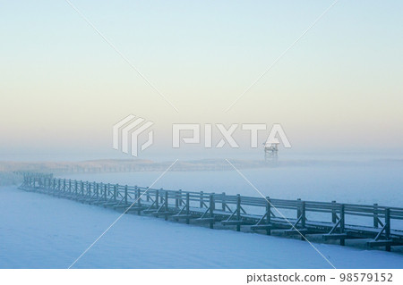 Winter view of a snow covered wooden boardwalk on a frozen lake leading to a bird watching tower 98579152