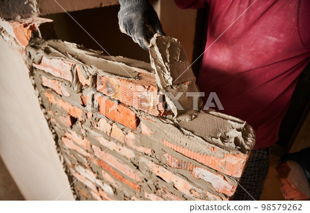 Close up of man hand in work glove laying brickwork in building under construction. Male worker applying cement mortar on bricks with trowel tool. Masonry construction concept. Close up of man hand in work glove laying brickwork in building under construction. Male worker applying cement mortar on bricks with trowel tool. Masonry construction concept. 98579262