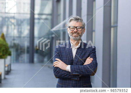 Portrait of mature gray-haired businessman, senior boss smiling and looking at camera with arms crossed, gray-haired man outside office building, successful investor and owner in business suit. Portrait of mature gray-haired businessman, senior boss smiling and looking at camera with arms crossed, gray-haired man outside office building, successful investor and owner in business suit. 98579352