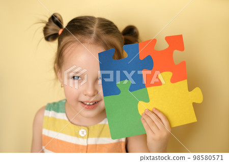 A beautiful attractive little blue-eyed girl smile cheerfully while closing eye holding puzzle of multicolored details in hand. Concept for Autism Day A beautiful attractive little blue-eyed girl smile cheerfully while closing eye holding puzzle of multicolored details in hand. Concept for Autism Day 98580571