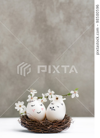 Happy Easter. White Easter eggs in wicker wooden nest on white table on gray background. View is straight. Blossoming branch of cherry with white flowers. Stylish spring template with copy space Happy Easter. White Easter eggs in wicker wooden nest on white table on gray background. View is straight. Blossoming branch of cherry with white flowers. Stylish spring template with copy space 98580596