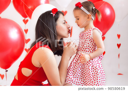 Mother and daughter with heart-shaped lollipops on a white background with decor for Valentine's Day. The woman's red dress and the daughter's white heart-shaped dress Mother and daughter with heart-shaped lollipops on a white background with decor for Valentine's Day. The woman's red dress and the daughter's white heart-shaped dress 98580619