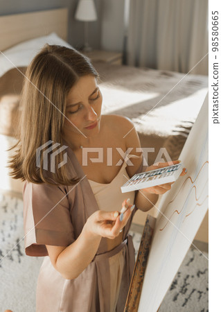 Young woman artist painting on canvas on the easel at home in bedroom - art and creativity concept Young woman artist painting on canvas on the easel at home in bedroom - art and creativity concept 98580665