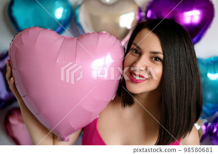 A young beautiful brunette woman holds a pink balloon in the shape of a heart in her hand, smiling sweetly. Valentine's day A young beautiful brunette woman holds a pink balloon in the shape of a heart in her hand, smiling sweetly. Valentine's day 98580681
