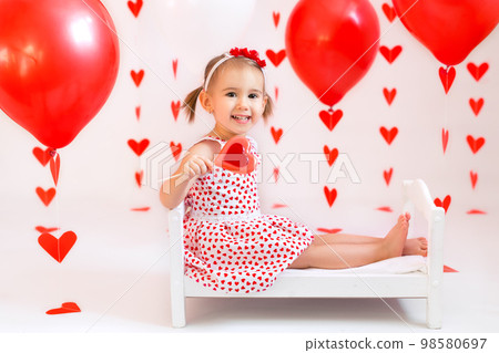 Baby holds red balls for Valentine's Day. A girl is eating a candy in the shape of a heart. A child in a white dress with hearts. Red hearts on a white background. 98580697