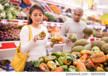 Positive woman picking ripe bell peppers at supermarket 98584030