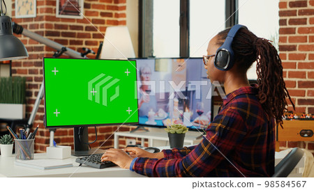 Female freelancer wearing headphones and using greenscreen on computer desktop, sitting at desk and working with isolated chroma key display. Looking at monitor with blank mock up. 98584567