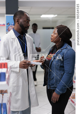 Pharmacy customer buying medication, pharmacy consultant recommending buyer pills, side view. African american woman talking with man pharmacist in drugstore aisle, all black team Pharmacy customer buying medication, pharmacy consultant recommending buyer pills, side view. African american woman talking with man pharmacist in drugstore aisle, all black team 98584585