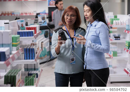 Asian women examining bottle of pills in pharmacy shop to buy medical supplies and health care products from drugstore shelves. Consultant helping client with pharmaceutical drugs. 98584702