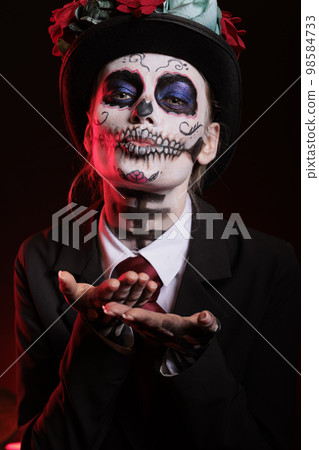 Woman doing kissy face with santa muerte make up, blowing air kisses at camera. Model wearing dios de los muertos make up and costume to celebrate mexican halloween holiday in studio. Woman doing kissy face with santa muerte make up, blowing air kisses at camera. Model wearing dios de los muertos make up and costume to celebrate mexican halloween holiday in studio. 98584733