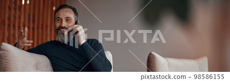 Smiling bearded man sitting in armchair and chatting on phone Smiling bearded man sitting in armchair and chatting on phone 98586155