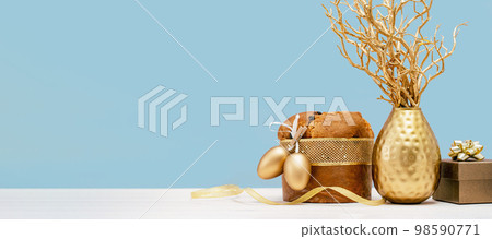 Happy Easter celebration banner. Italian Easter Decorated Panettone and golden eggs. tree branches in a vase and a gift box on white table. Wide banner on blue. 98590771