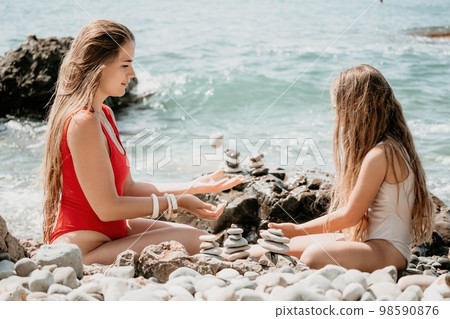 Woman with daughter bilds stones pyramid on seashore on a sunny day on the blue sea background. Happy family holidays. Pebble beach, calm sea. Concept of happy vacation on the sea, meditation, spa 98590876