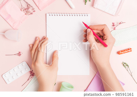 Lady with red felt-tip pen over notepad on pink background with pills and sanitary napkin 98591161