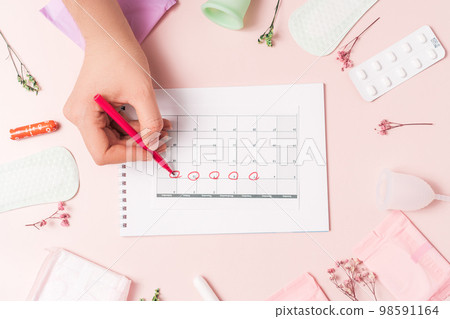 Lady marks with red felt-tip pen in the calendar the menstrual cycle on pink background with pills and sanitary napkin 98591164