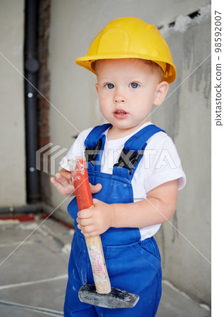 Adorable child construction worker holding hammer building repair tool and looking at camera. Cute kid wearing safety helmet and work overalls while posing at home during renovation. 98592007