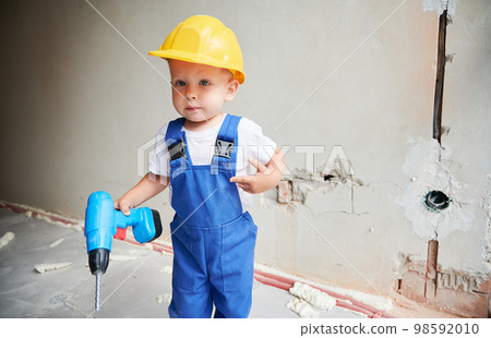 Child with toy construction tool for repair works standing against the wall in apartment under renovation. Kid in safety helmet and work overalls holding electric drill and looking at camera. Child with toy construction tool for repair works standing against the wall in apartment under renovation. Kid in safety helmet and work overalls holding electric drill and looking at camera. 98592010