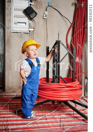 Adorable little boy standing near underfloor heating pipes. Cute kid plumber wearing work overalls and safety helmet while posing in apartment under renovation. Adorable little boy standing near underfloor heating pipes. Cute kid plumber wearing work overalls and safety helmet while posing in apartment under renovation. 98592011
